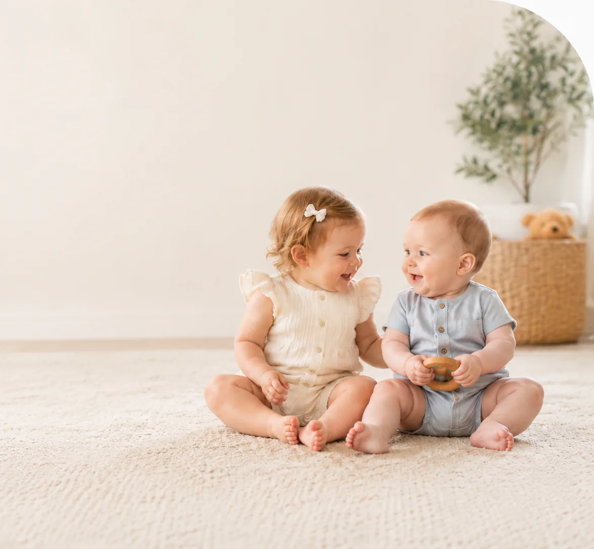 Two babies sitting together on a soft rug, smiling at each other